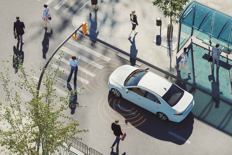 Driverless car with orange environmental sensors approaching a pedestrian crossing. A number of people are crossing the road.