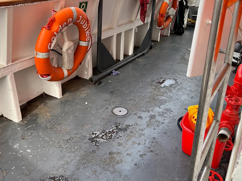 The deck of a boat with water on it. A bright orange life buoy is in the background and a red bucket