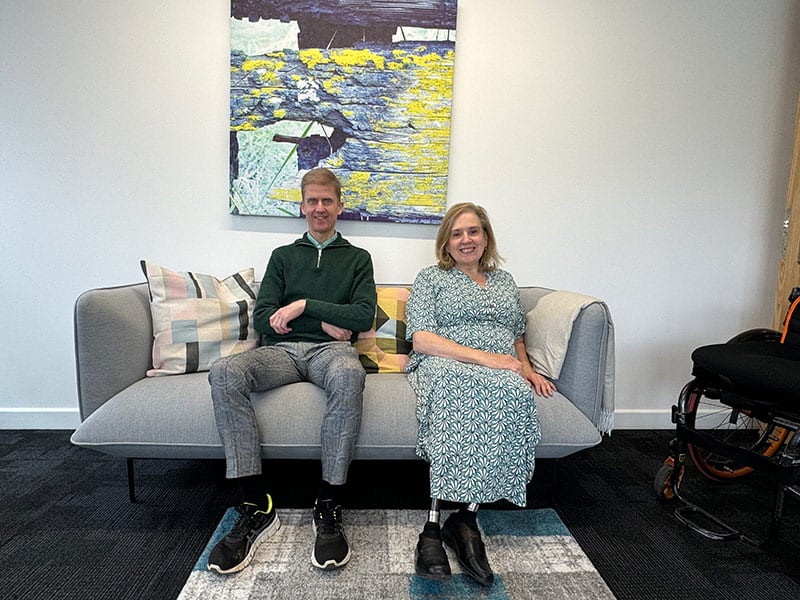 Liam and Tracey Proudlock sat on a grey sofa in front of a colourful painting. Both are smiling