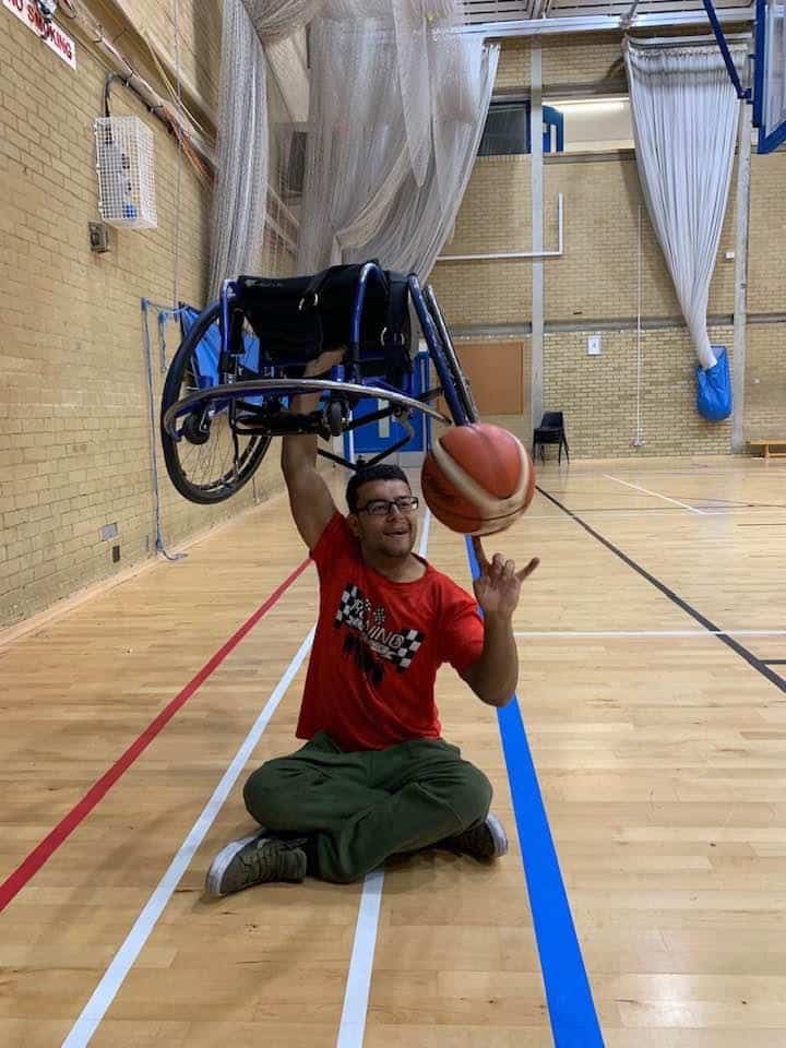 A man sat on the floor holding his wheelchair above his head with one hand and spinning a basketball in the other.
