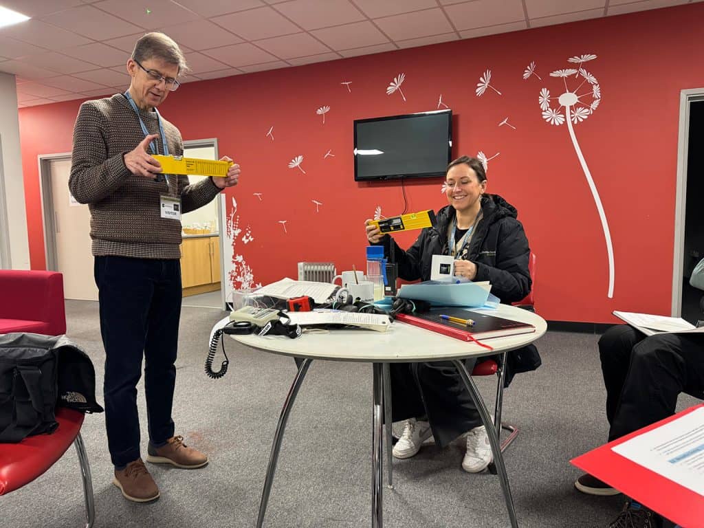 Liam is standing up and holding a yellow spirit yellow. There is a desk in front of him where a woman is sat. She is also holding a spirit level and smiling. The room is painted red with white flowers.