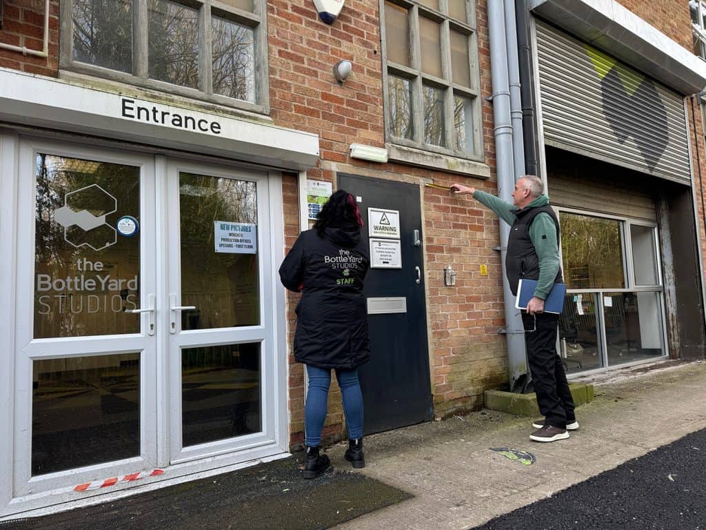 The BottleYard Studios' entrance. The double door is white with the word Entrance written above. A woman and man stand to the right of the door and are measuring a black door with a warning sign on it. More windows and a retractable ridged door with a grey and yellow mark can be seen in the background.