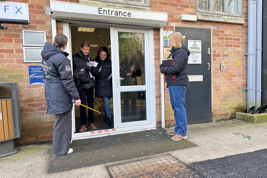 The BottleYard Studios' entrance. The double door is white with the word Entrance written above. The left side of the door is open and a man and woman stand in it, smiling. There is also a woman just outside the door on the left holding a tape measure and a woman on the right holding a book.