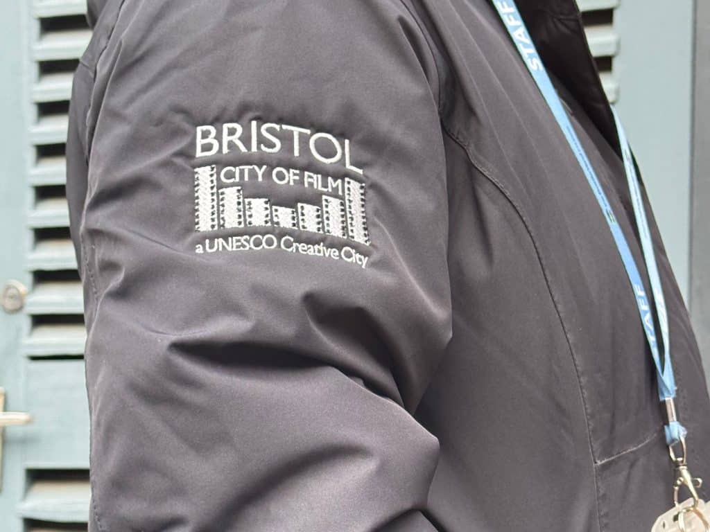 A white logo which says Bristol City of Film a Unesco Creative City with pieces of film. The logo is on the sleeve of a grey jacket worn by someone with a blue lanyard around their neck.