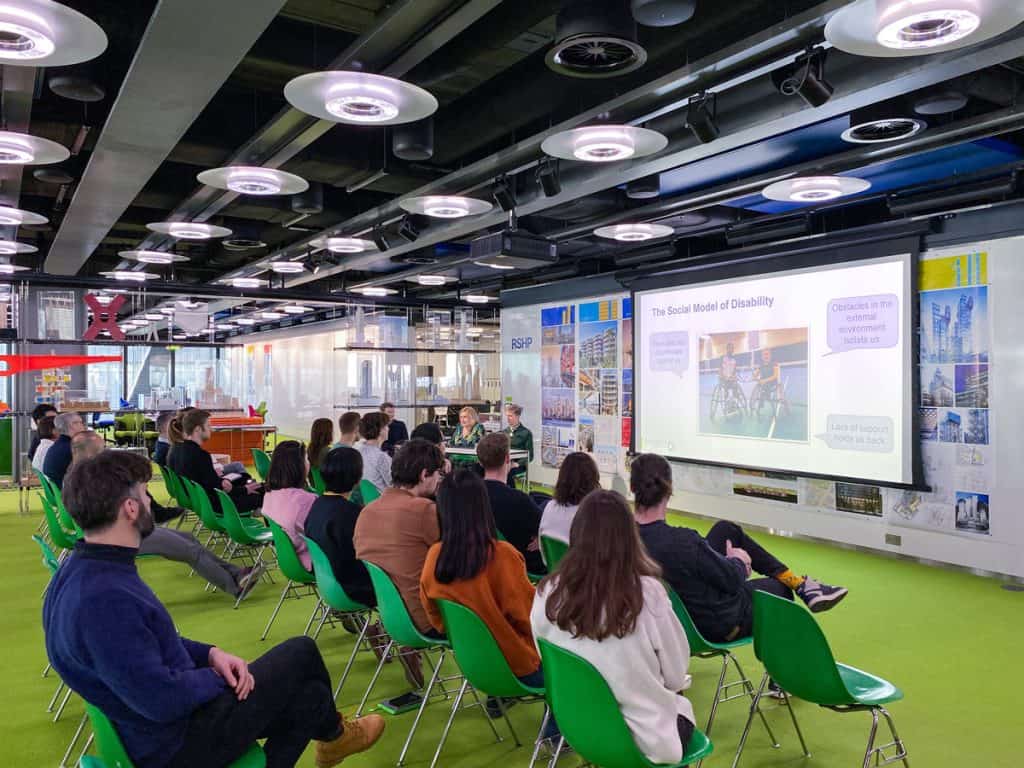 A group of people sat on green chairs in a conference room with their backs to the camera. They are looking at a giant screen which has the words The Social Model of Disability in bold. Three people sit to the lefthand side of the screen behind a table.