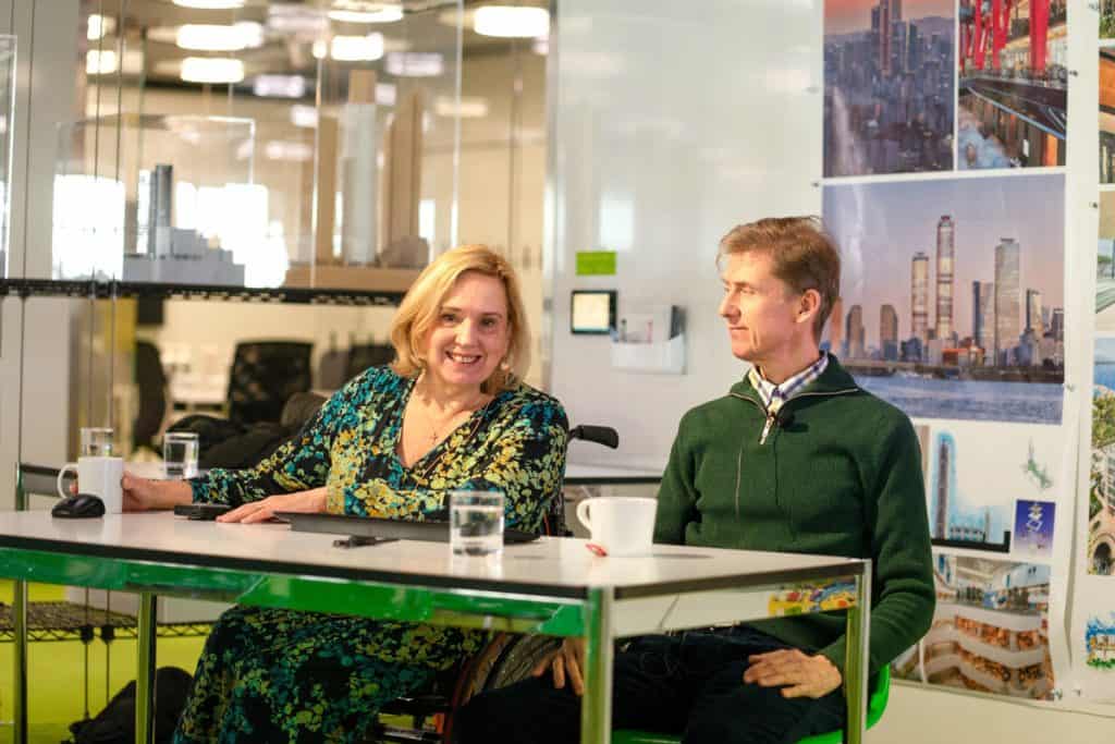 Tracey and Liam sat at a desk. Tracey is smiling at the camera and Liam is looking at Tracey. There are two cups and a glass on the desk. In the background there is a banner stand showing image of cities and a glass wall with reflections.