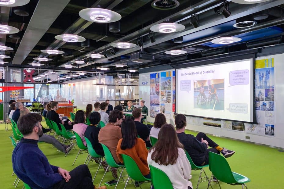 A group of people sat on green chairs in a conference room with their backs to the camera. They are looking at a giant screen which has the words The Social Model of Disability in bold. Three people sit to the lefthand side of the screen behind a table.