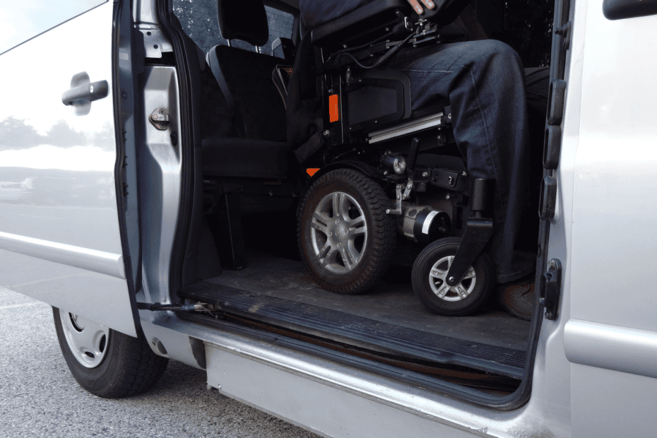 A person in a power wheelchair enters a wheelchair-accessible van using a lowered floor and ramp system. The side door of the silver van is open, showing the accessible interior.