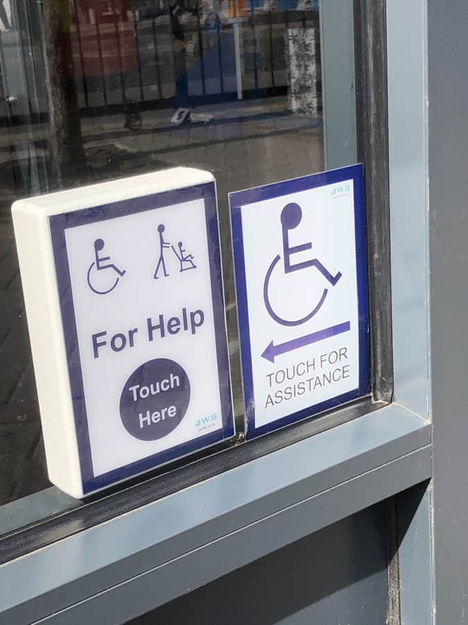 A photo of a window with a grey frame. It has two signs. One has a picture of a wheelchair user and someone pushing a pushchair and has the words For Help and then Touch Here in a big blue circle. The other sign has a picture of a wheelchair user, an arrow pointing to the left and the words 'Touch for Assistance'. 