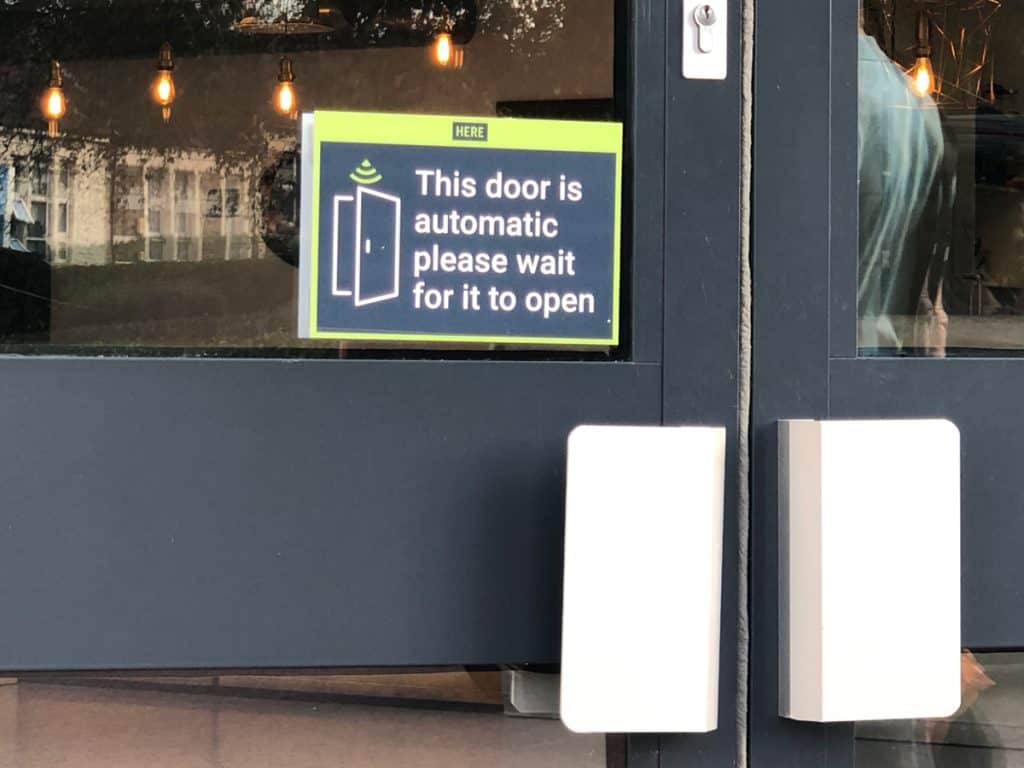 A photo of a pair of grey glass doors with chrome handles. The is a sign in dark blue with a green border which says "This door is automatic please wait for it to open". 