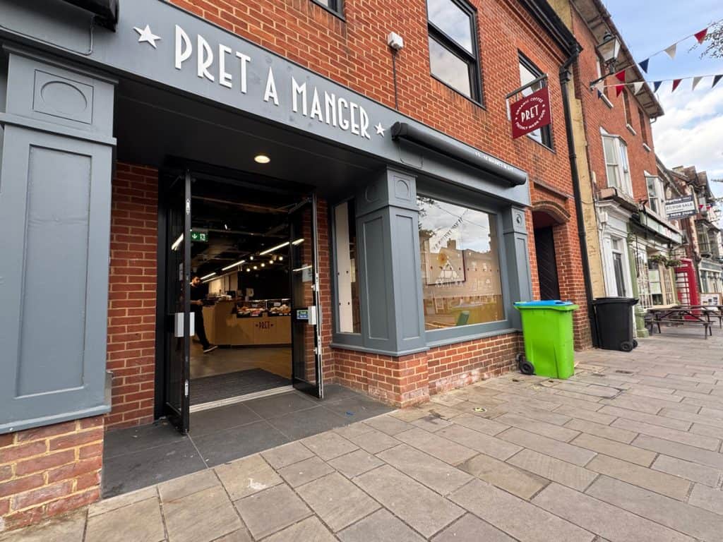 A photo of a Pret A Manger store front. The exterior is grey and the doors are open showing good level access into the store. The store is in a high street. 