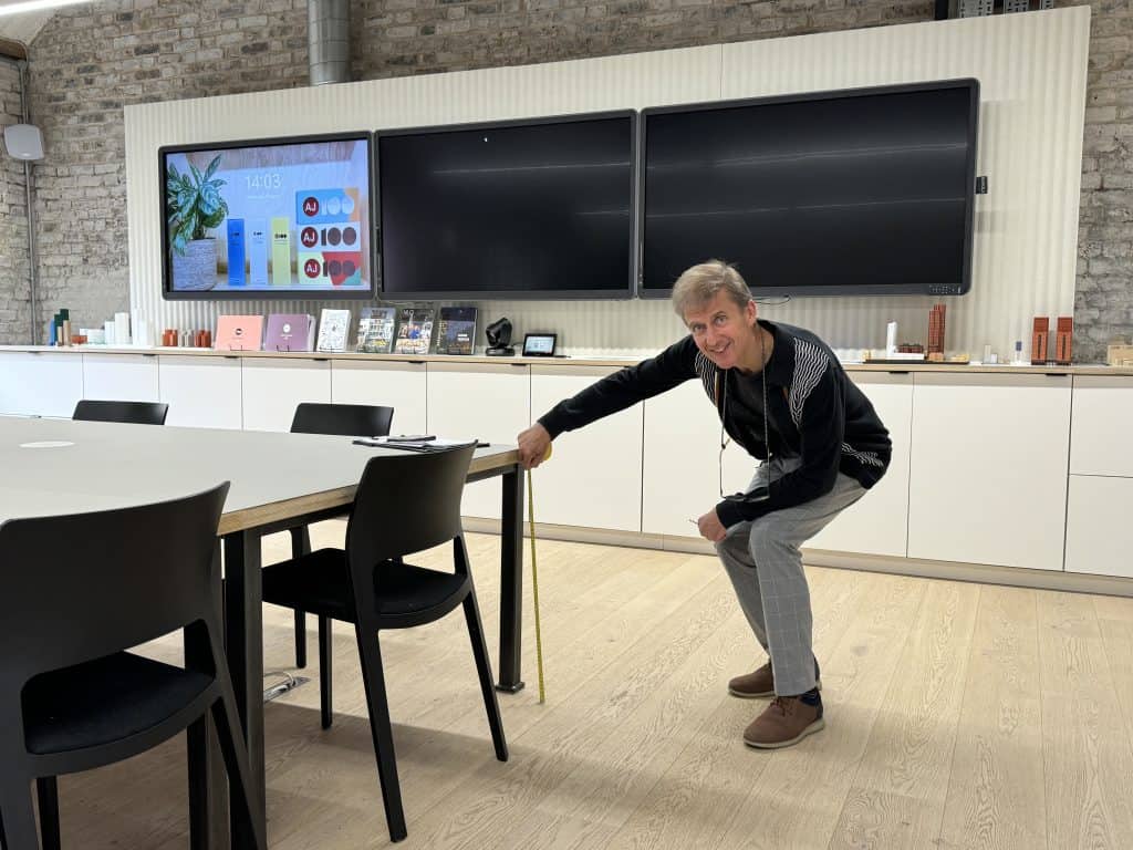 An office with white cupboards and TV screens on the wall in the background. In the front is a table with black chairs. A white man is bending down and measuring the height of the table with a tape measure. He is wearing a black jumper with white stripes down the arms and grey checked trousers. He is smiling at the camera.