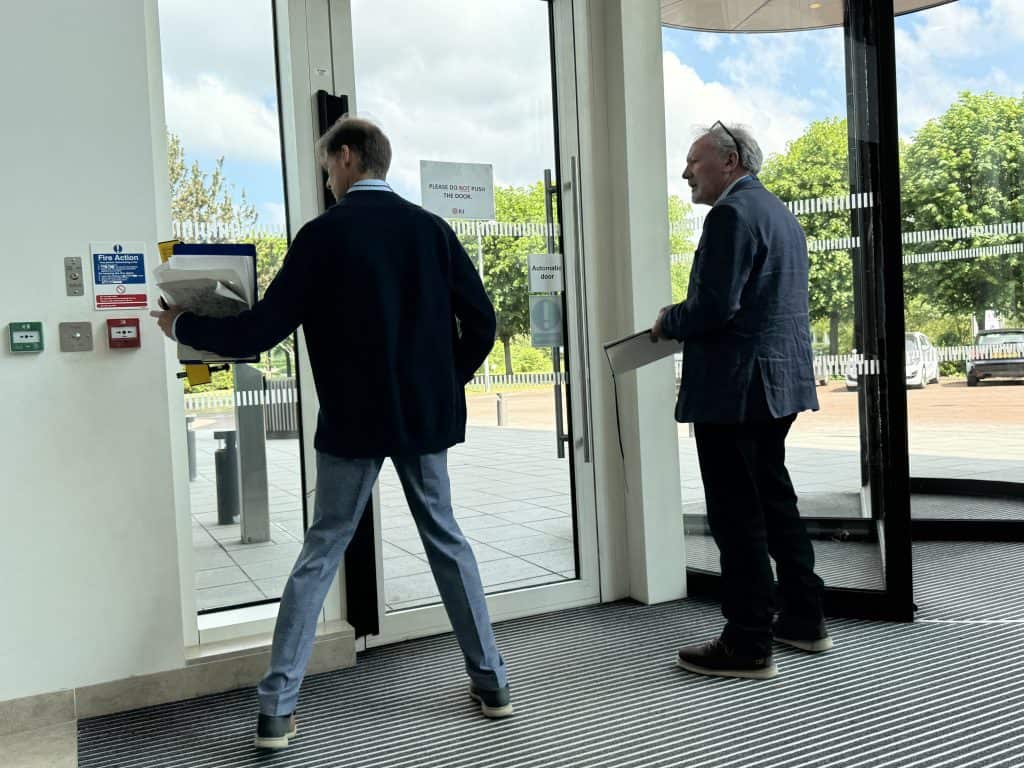A photo of doors to an office building from the inside. On the left is a push open door and on the right a revolving door. There is a white man on the left with his back to the camera who is pointing at the fire alarm button. He is wearing a black jumper, jeans and is holding a file of papers in his hand. There is another white man to the right who is watching what's going on. He has glasses on his head, is wearing a blue jacket and black trousers and has a file in his hand. Outside the doors you can see trees and two parked cars.