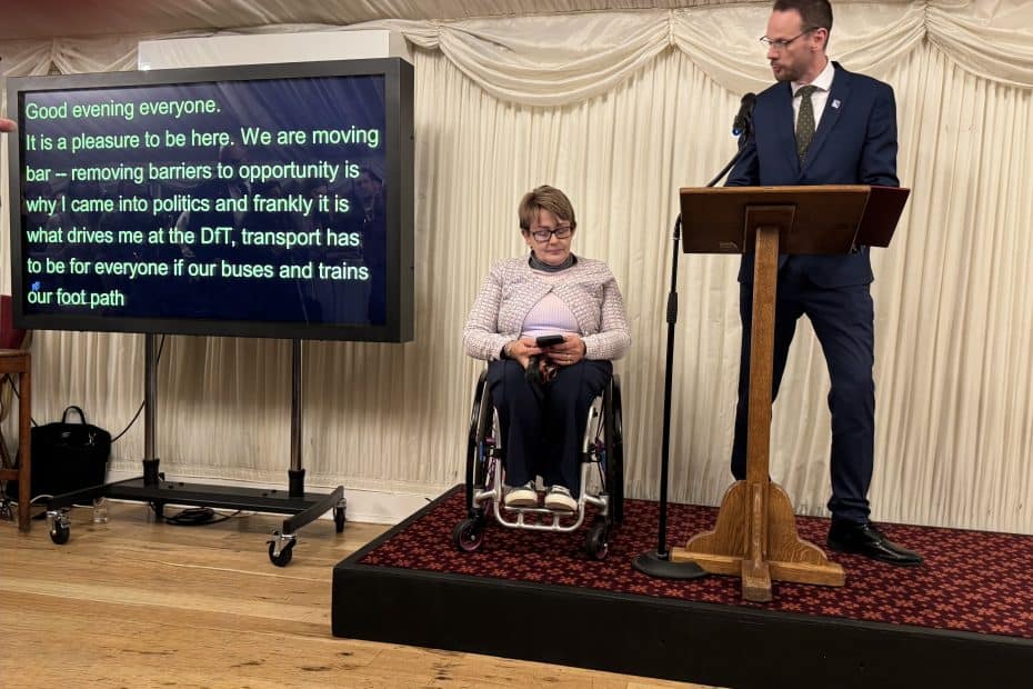 Simon Lightwood MP stands at the lectern speaking on a stage. Next to him is Baroness Tanni grey-Thompson. To the left, a large screen displays live captions of the speech. The setting is the indoor reception of Cholmondeley Room at the House of Lords.