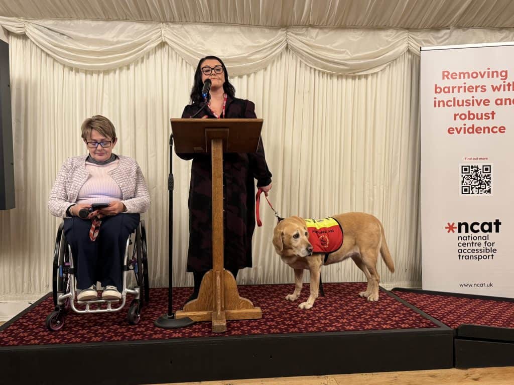 Emma Partlow stands at the lectern speaking into a microphone on a small stage with an assistance dog wearing a red working vest. Beside her is Baroness Tanni Grey-Thompson. A banner for the National Centre for Accessible Transport is visible in the background.