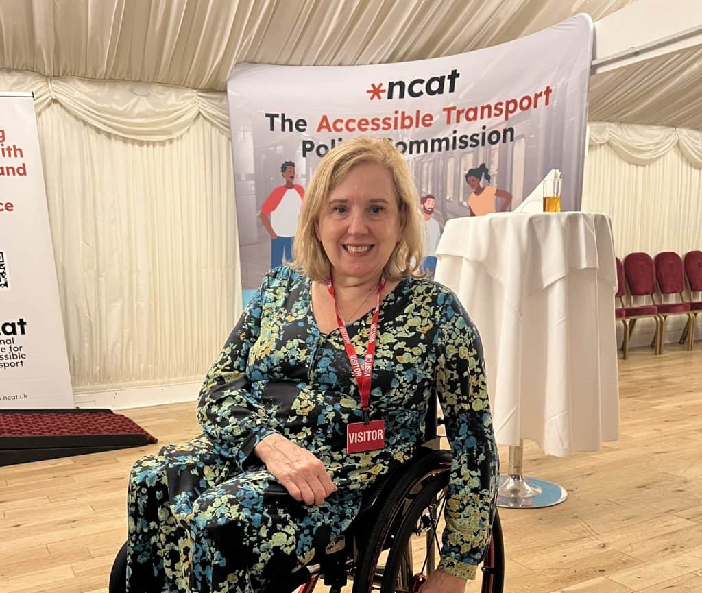 Tracey Proudlock posing in the reception room of Cholmondeley Room at the House of Lords. Behind her, a banner reads “The Accessible Transport Policy Commission” with illustrated figures representing diverse transport users.