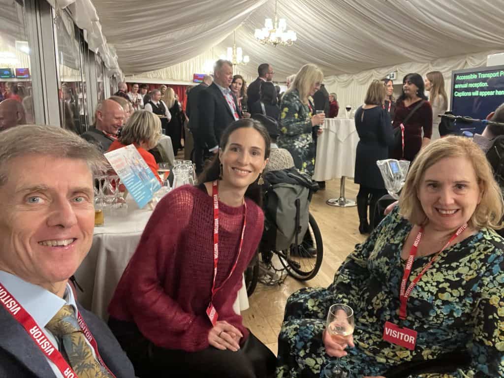 From left to right, Liam Proudlock, Regina Bortoni and Tracey Proudlock wearing visitor badges pose for a photo at the indoor reception of Cholmondeley Room at the House of Lords. Other attendees, tables, and chandeliers are visible in the background.