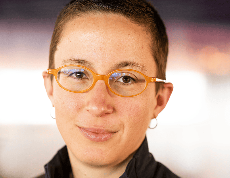 A portrait photograph of Itxaso Markiegi. They have short dark hair and are looking straight at the camera. They are wearing yellow framed glasses and a black top.