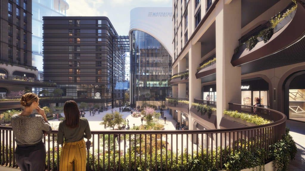 Artist's impression of London Motorsports campus. There are railings at the front of the image with two people stood looking over a plaza which features trees and shrubs and people walking. To the left is a building with balconies festooned with plants. At the far end is a glass building.