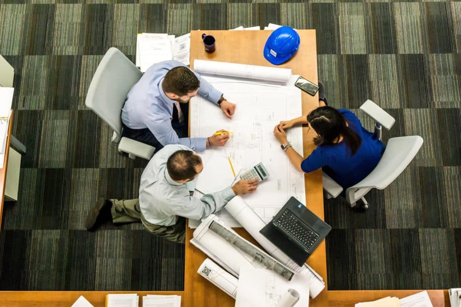A picture taken from above of three people sat around the table. On the table are architects drawings, a laptop and a blue hard hat. One man is holding a calculator and a yellow marker pen.