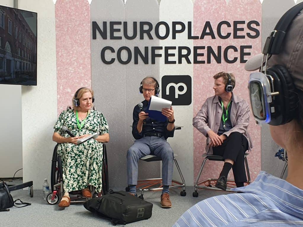 A photo taken at the Neuroplaces Conference. Tracey Proudlock, Liam Proudlock and Mark Rintoul are sat on the stage. Liam is looking at a piece of paper and Mark is looking at him. The background of the stage is grey, pink and white stripped and says Neuroplaces Conference in large black letters. Underneath is the Neuroplaces logo.