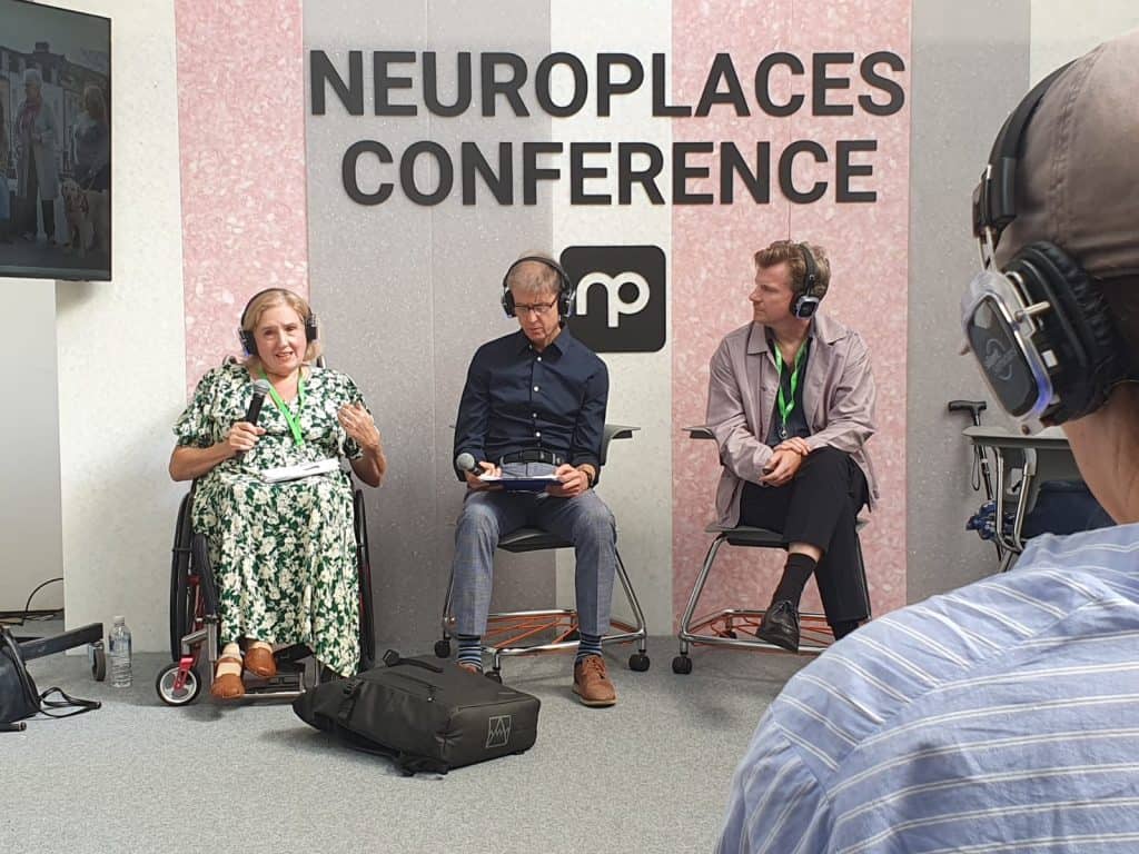A photo taken at the Neuroplaces Conference. Tracey Proudlock, Liam Proudlock and Mark Rintoul are sat on the stage. Liam is looking at a piece of paper on his lap and Mark is looking at Tracey who is talking. The background of the stage is grey, pink and white stripped and says Neuroplaces Conference in large black letters. Underneath is the Neuroplaces logo.