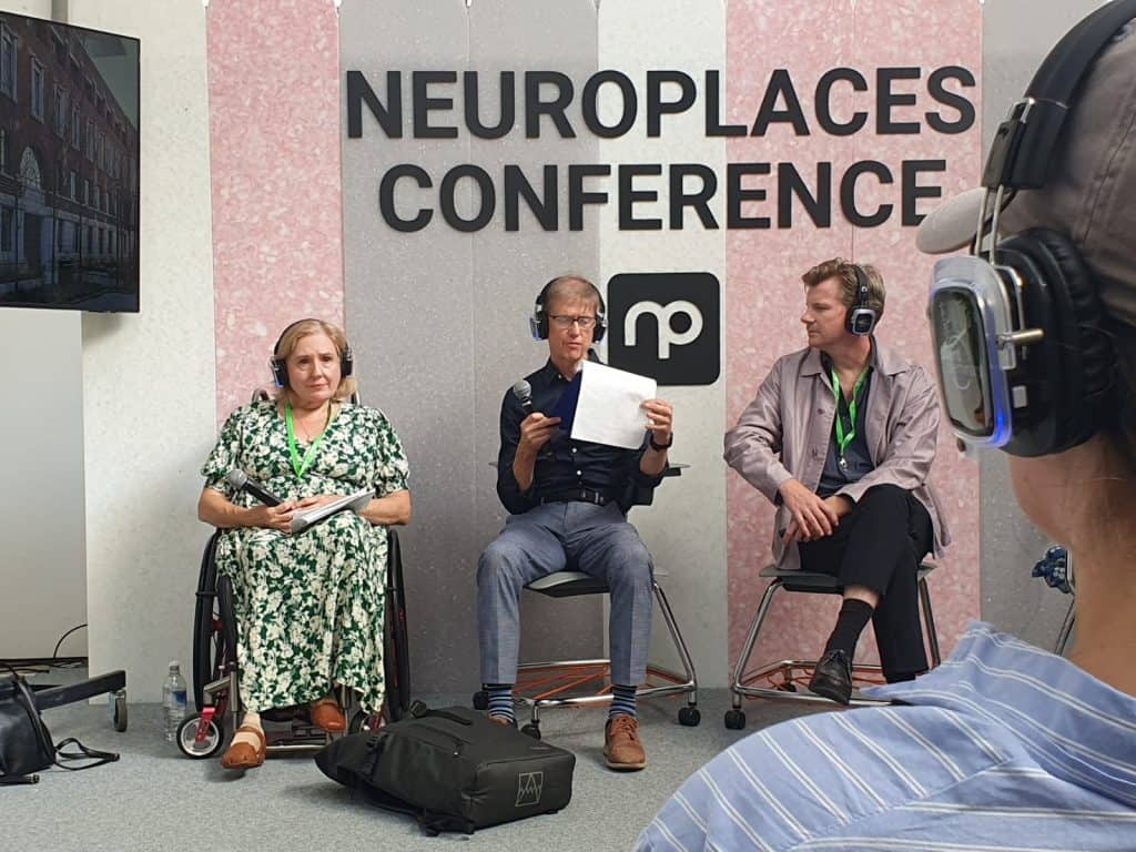 A photo taken at the Neuroplaces Conference. Tracey Proudlock, Liam Proudlock and Mark Rintoul are sat on the stage. Liam is looking at a piece of paper and Mark is looking at him. The background of the stage is grey, pink and white stripped and says Neuroplaces Conference in large black letters. Underneath is the Neuroplaces logo.