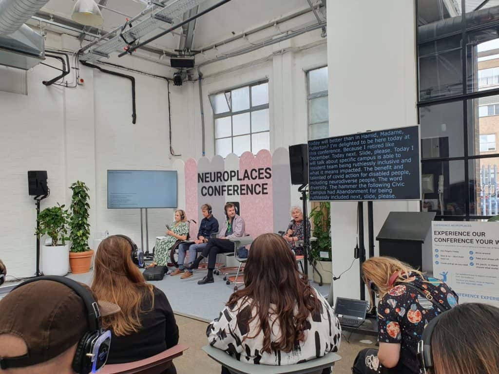 A photo taken at the Neuroplaces Conference. It shows four people on a stage with a TV screen and a screen showing words. In the foreground is a group of people watching the stage.